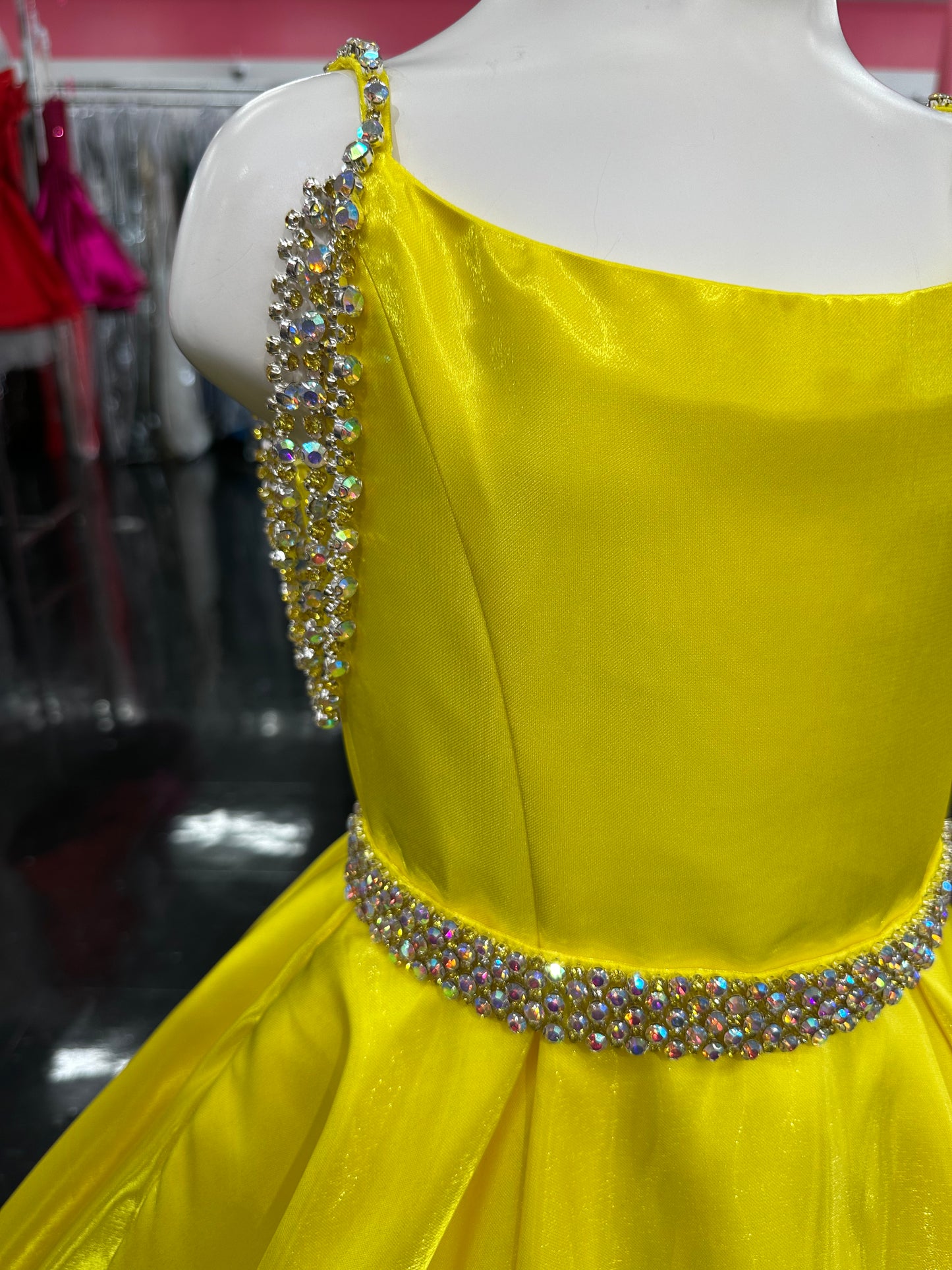 Ashley Lauren 8333, a stunning satin A-line gown designed for young competitors. This sophisticated dress features shimmering fabric that catches the light beautifully, complemented by elegant crystal-embellished off-the-shoulder straps that frame the neckline with glamour. The coordinating belt cinches the waist for a polished silhouette, while practical hidden pockets add functionality without compromising style. The flowing A-line skirt creates graceful movement on stage, ensuring your daughter commands