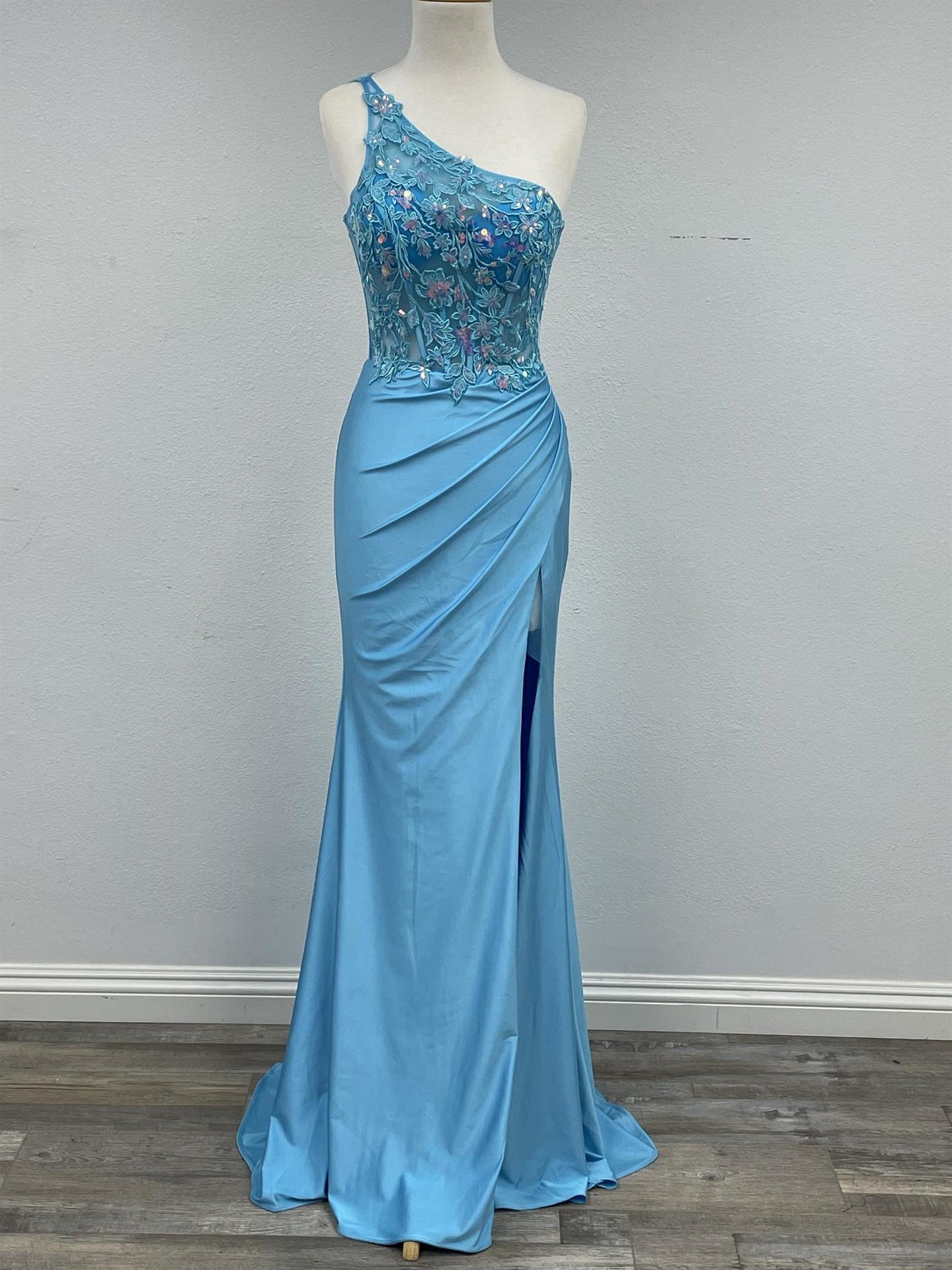 This elegant Nox Anabel A1317 dress features a sheer one shoulder design with a corset bodice and a sequin lace overlay. The fitted silhouette flatters the figure, while the slit adds a touch of sophistication. Perfect for prom or any formal occasion, this gown is sure to make a statement.