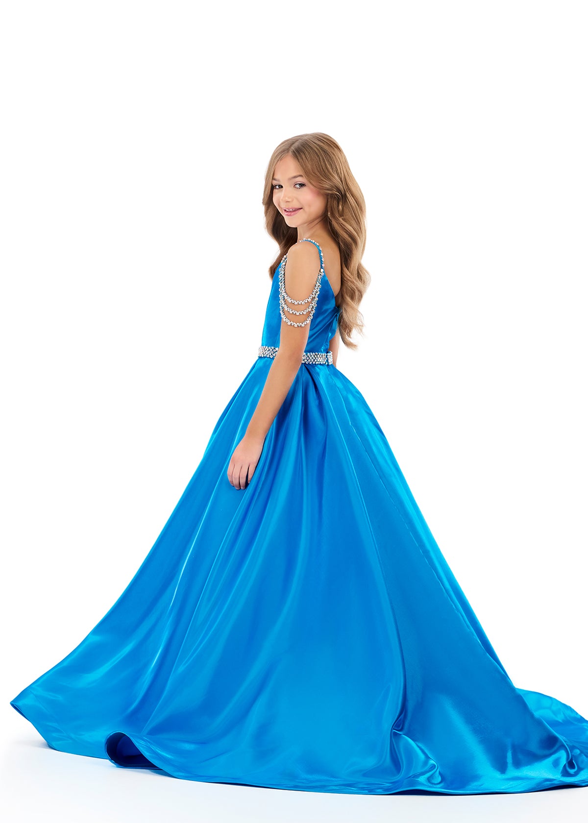 Ashley Lauren 8333, a stunning satin A-line gown designed for young competitors. This sophisticated dress features shimmering fabric that catches the light beautifully, complemented by elegant crystal-embellished off-the-shoulder straps that frame the neckline with glamour. The coordinating belt cinches the waist for a polished silhouette, while practical hidden pockets add functionality without compromising style. The flowing A-line skirt creates graceful movement on stage, ensuring your daughter commands 