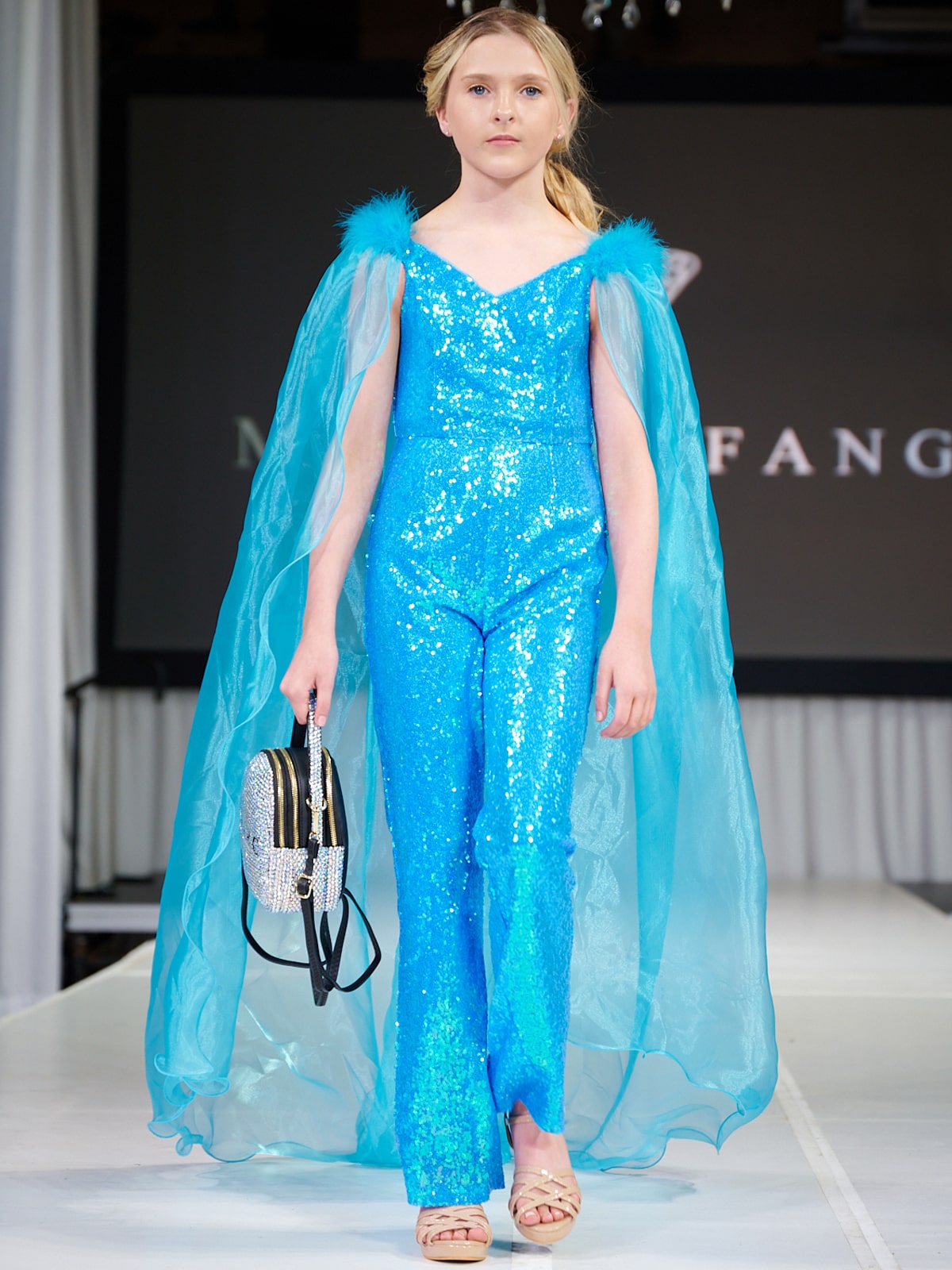 Marc Defang 8099K is a KIDS long pageant formal wear jumpsuit that is made of sequins and has off the shoulder straps.  The straps are attached to a cape with feathers at the shoulders.  Wow the crowd at your next pageant.  Available colors:  Pink, Turquoise, White, Black, Royal