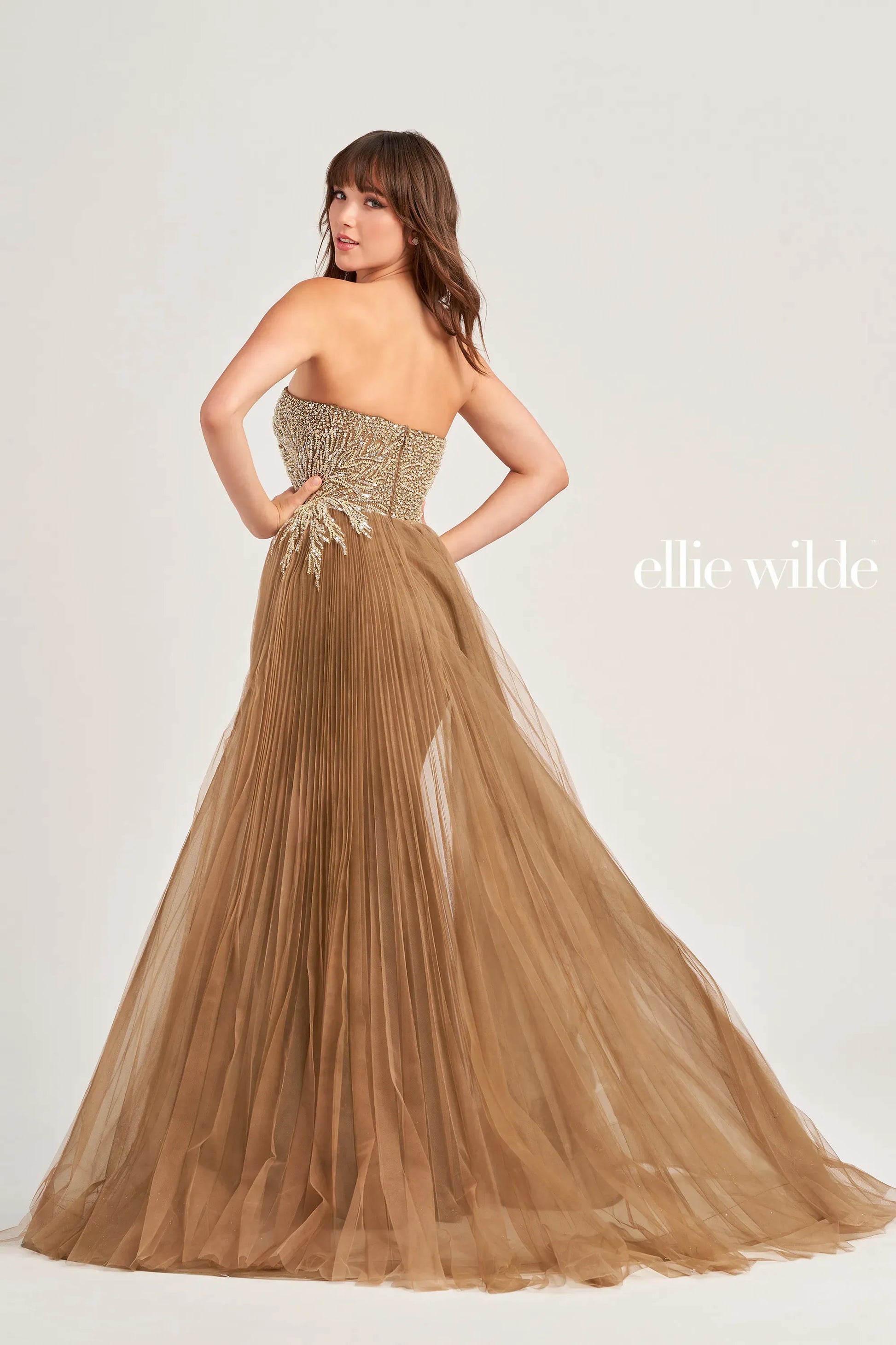 Elevate your formal look with the Ellie Wilde EW35085 Strapless Crystal Pageant Dress. Crafted with a high slit and pleated detachable overskirt, this gown exudes elegance and sophistication. The strapless design and crystal embellishments add a touch of glamour, perfect for any special occasion.  Sizes: 00-16  Colors: Coffee, Emerald