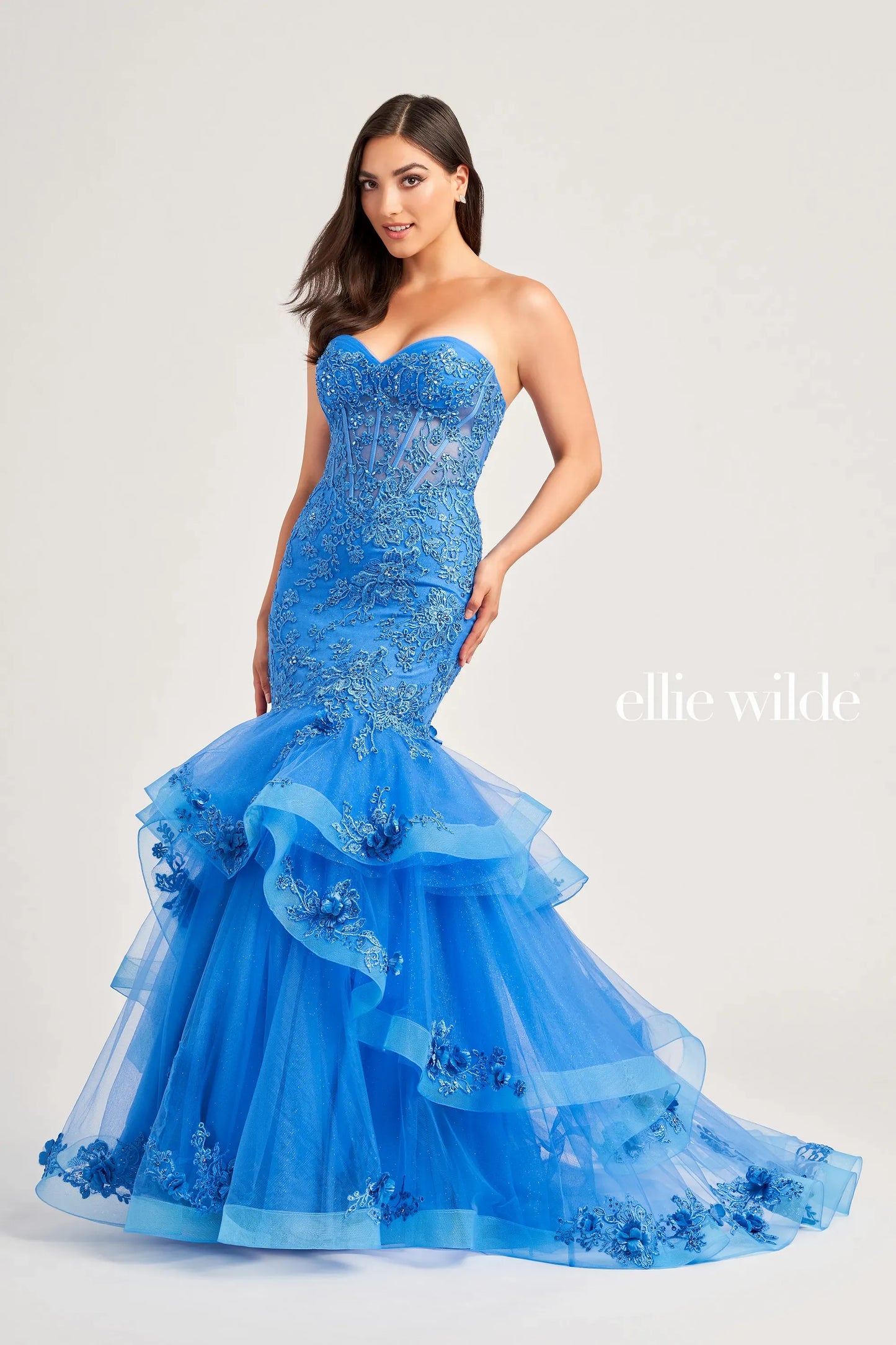 Get ready to make a statement in this Ellie Wilde EW35239 mermaid prom dress. The sheer corset lace bodice is adorned with embroidered and glitter tulle, while the ruffle layer skirt shimmers with stone accents and 3D flowers. You'll be the belle of the ball in this stunning dress.  Sizes: 00-16  Colors: Ocean Blue, Lilac, Red