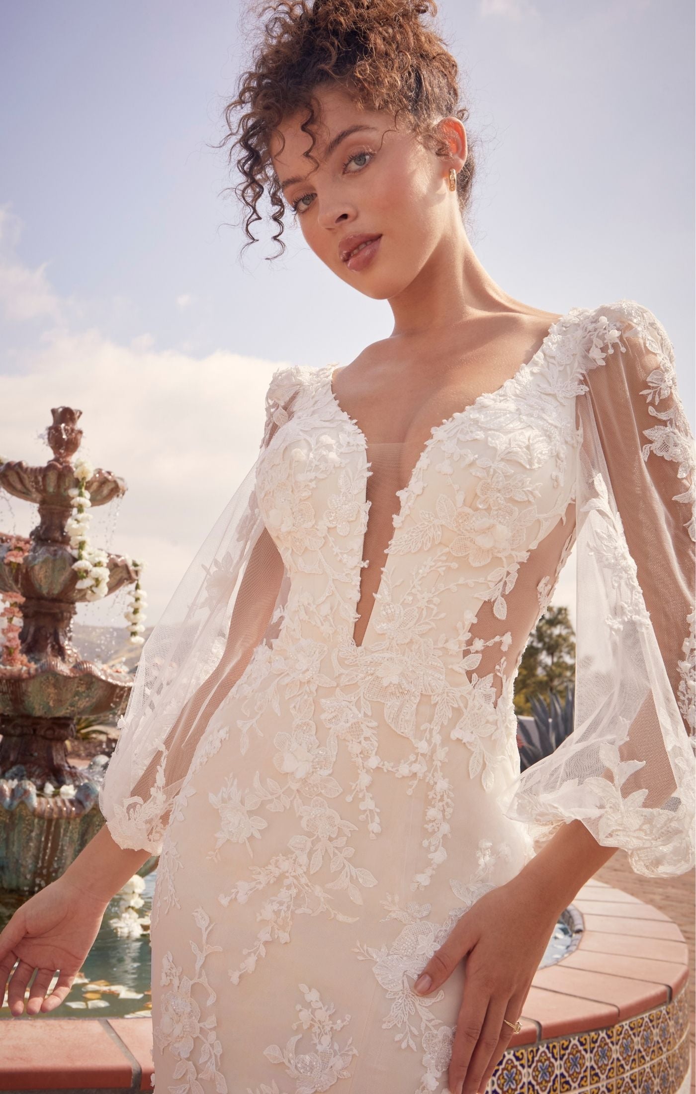 Casablanca Beloved BL434 Mermaid Sheer Long Sleeve V-Neck Open Back Lace Floral Tulle Train Wedding Gown. Undeniably feminine, Montie is the perfect mix between romantic floral lace motifs and figure flattering silhouettes. Perfect for the outgoing yet romantic bride, Montie’s plunging V neckline and curve sculpt lining bodice is contrasted with delicate feminine floral beaded & sequined lace, met with illusion balloon sleeves.
