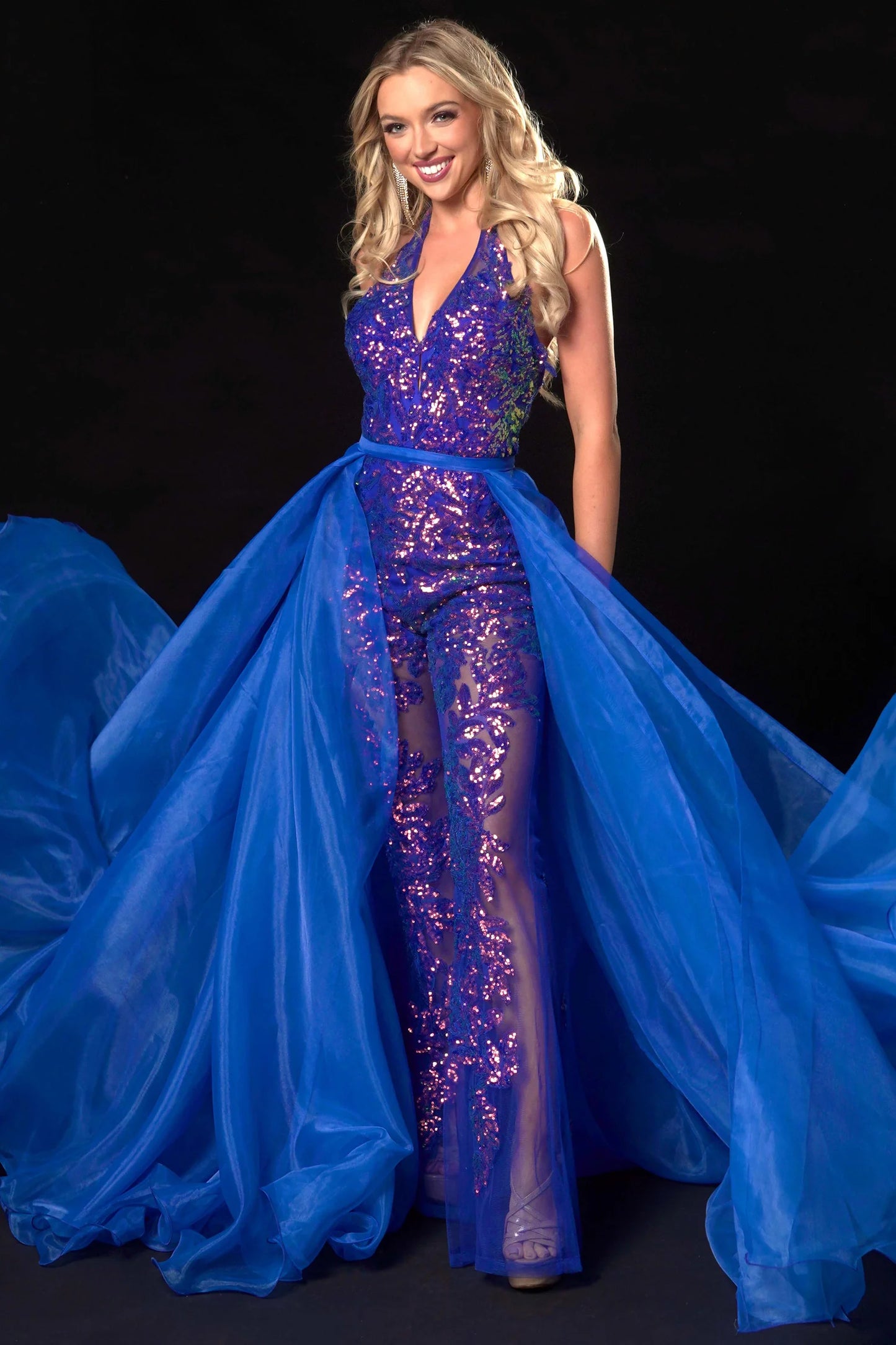 Ava Presley 37381 is crafted with a long, sheer sequin halter jumpsuit, detachable overskirt, V-neck backless design, and a hint of fun fashion. The result is a luxurious, glamorous look perfect for pageants.  Sizes: 0-16  Colors: Royal, Fuchsia, Iridescent Orange