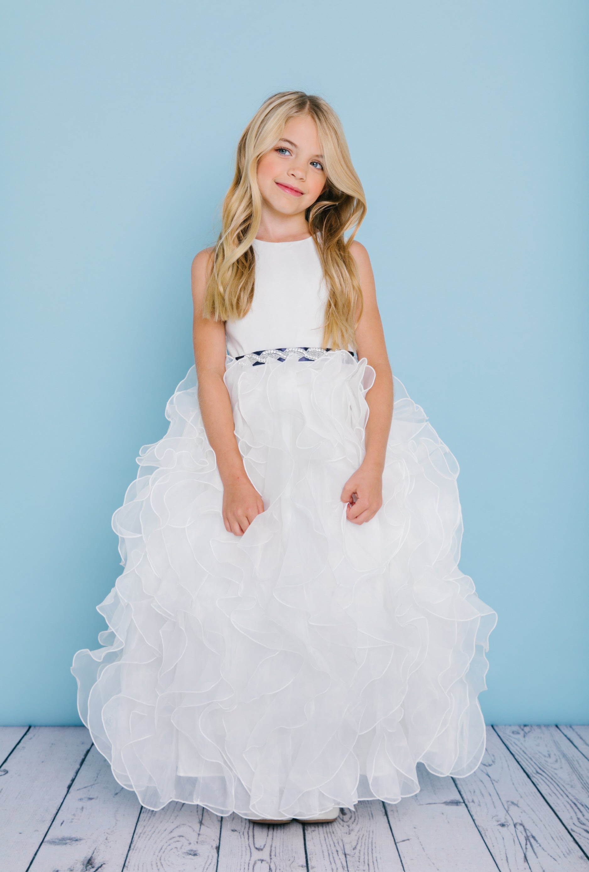 Rosebud Fashions Style 5122 is a ball gown with a satin bodice and organza ruffled skirt. The waist is accented by a bling ribbon that ties to a bow in the back. Satin buttons cover the zipper. Flower Girl Dress * First Communion Gown.  Available Sizes: 2,4,6,8,10,12  Available Colors: Ivory, White  Fabric is Satin and Organza