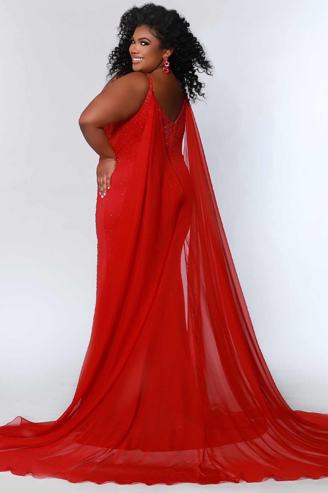 Johnathan Kayne for Sydney's Closet JK2218 Maverick Plus Sized Prom, Pageant and Formal Evening Wear Dress with Cape Buckle up because this gown is a hot one! The stretch heavy knit in this slim fit princess line silhouette and deep v-neckline makes showing off your curves a power move! Sexy from head to toe in Deep Red, Hot Coral, and White Ice, you'll catch the light with sparkling stones throughout the gown. Detachable chiffon scarves are the final winning moment and will fly as you strut!﻿