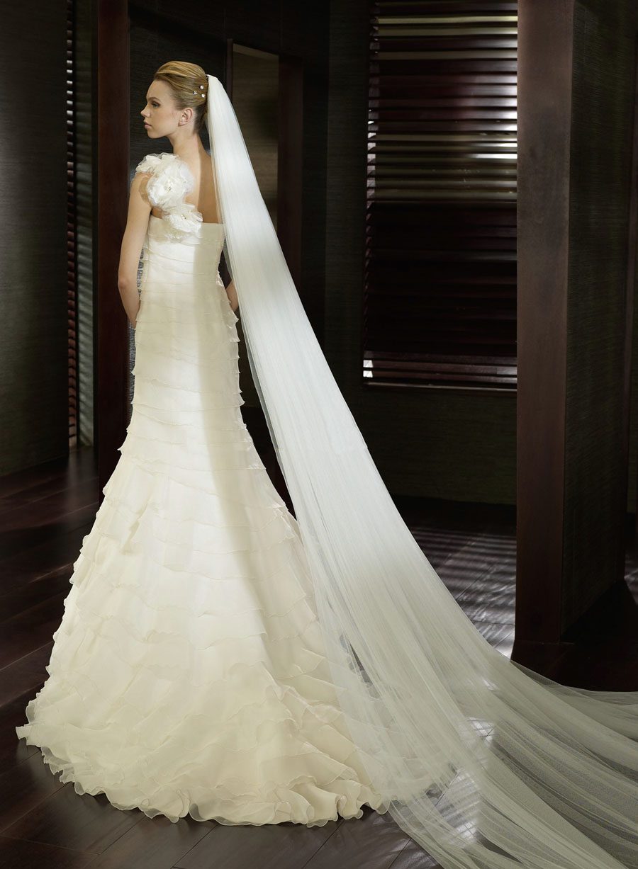 San Patrick Collection by Pronovias. San Patrick Eden is an A-line gown of layered Paris Gauze with a one shoulder neckline with a large flower on the shoulder strap. The layers on the skirt part are ruffled. Train in back.  Available in Size 10 in Off White