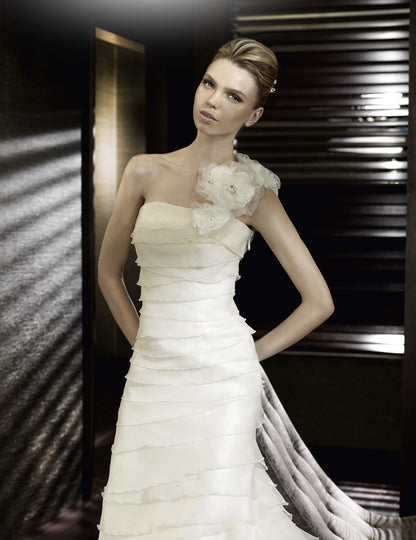 San Patrick Collection by Pronovias. San Patrick Eden is an A-line gown of layered Paris Gauze with a one shoulder neckline with a large flower on the shoulder strap. The layers on the skirt part are ruffled. Train in back.  Available in Size 10 in Off White