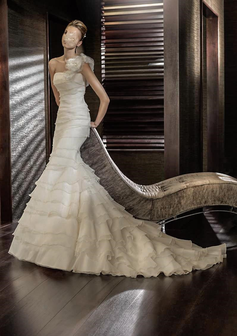 San Patrick Collection by Pronovias. San Patrick Eden is an A-line gown of layered Paris Gauze with a one shoulder neckline with a large flower on the shoulder strap. The layers on the skirt part are ruffled. Train in back.  Available in Size 10 in Off White