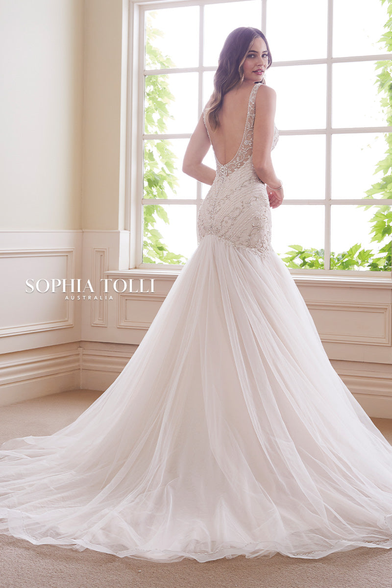 Sophia Tolli Y21811 KYANITE This Sophia Tolli Y 21811 Kyanite trumpet-style wedding gown has an intricately beaded bodice, with illusion straps edging the V-neckline and open scoop back. The gathered tulle skirt begins on the drop waist, and flows to a beaded horsehair hem and chapel train. Diamante buttons trim the back zipper closure. 